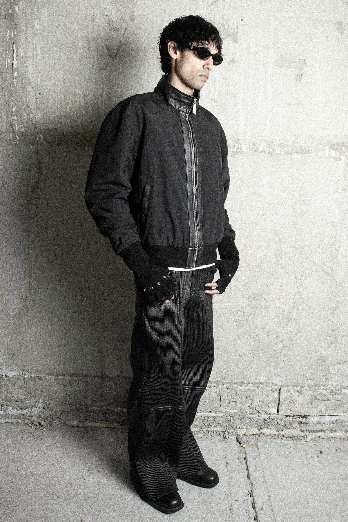 A man wearing a black bomber jacket, black pants, black fingerless gloves, and black sunglasses, standing against a textured concrete wall.